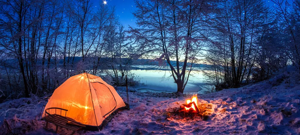 Cozy winter camping scene with a glowing tent and campfire by a frozen lake under stars.