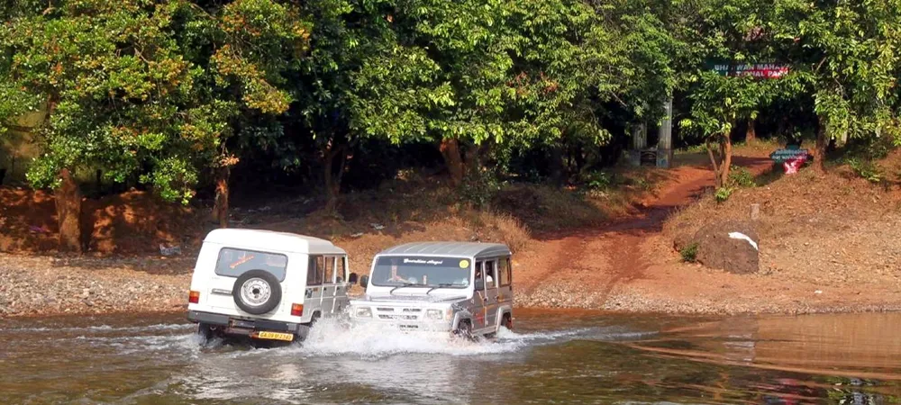 Two SUVs driving through shallow water in a forest, with a sign for a national park in the background.
