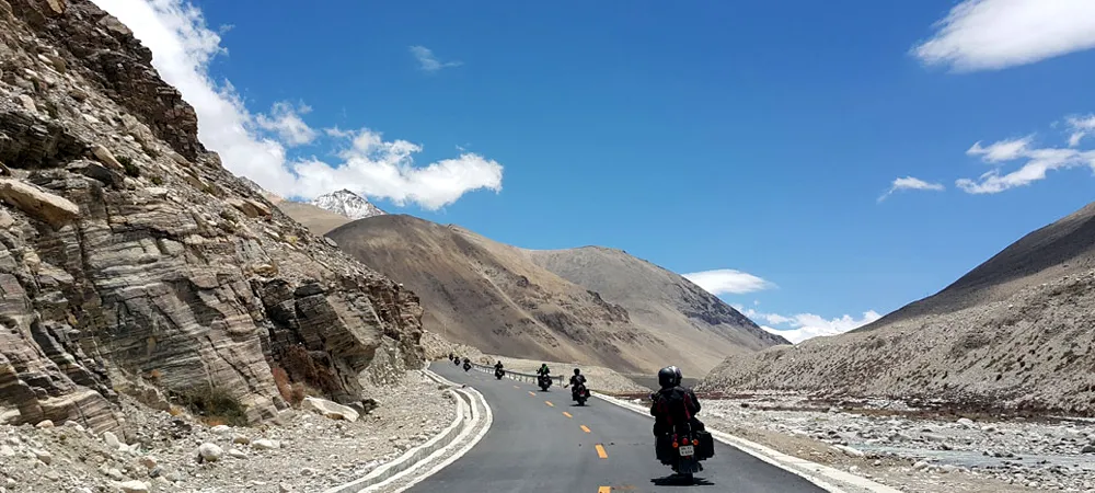 Motorcyclists ride on a scenic mountain road in a remote, arid landscape under a blue sky.